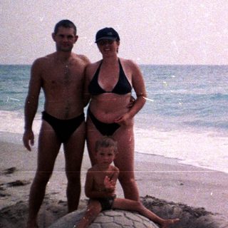 Family poses near a sand turtle sculpture on a beach.