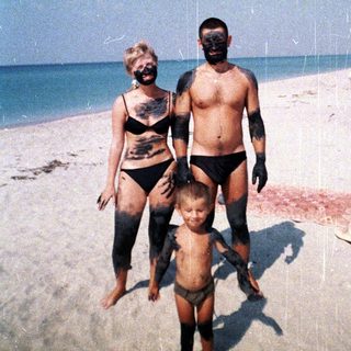 Family covered in dark mud stands on a beach.