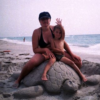 Woman and child sit atop a large sand turtle sculpture.