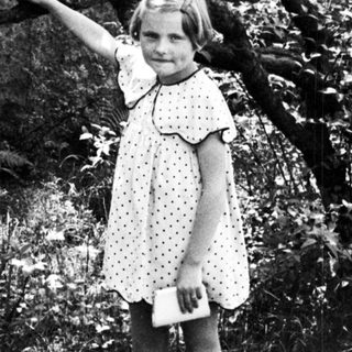 Girl in dress stands on path, reaching toward tree.