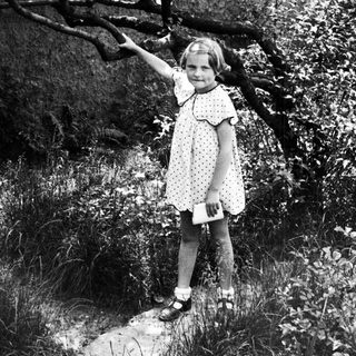 Girl in dress stands on path, reaching toward tree.