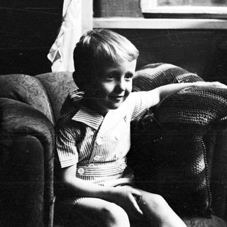 Here's a brief summary: Young boy seated on a patterned armchair, looking toward the viewer.