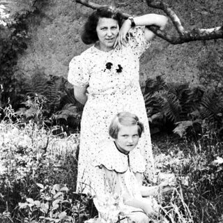 Woman and child stand in a garden, foliage visible.