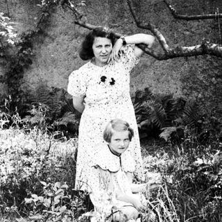 Woman and child stand in a garden, foliage visible.