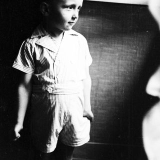 Here's a brief summary of the image: Young boy in striped shorts stands near a window.