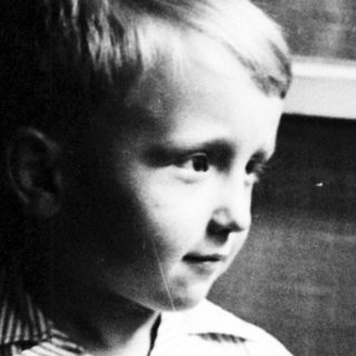 Here's a brief summary of the image: Young boy in striped shorts stands near a window.