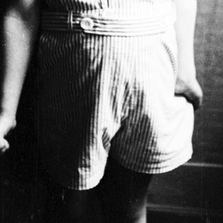 Here's a brief summary of the image: Young boy in striped shorts stands near a window.