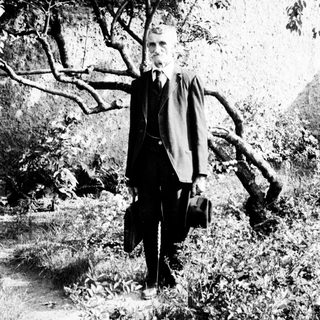 Man in suit stands outdoors, holding a hat and briefcase.