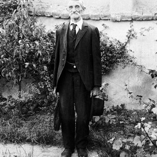 Elderly man in suit stands outdoors with a briefcase.