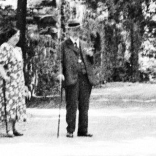 Man and woman stand under stone archway, trees beyond.