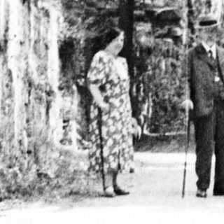 Man and woman stand under stone archway, trees beyond.
