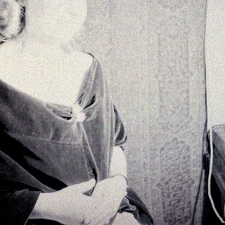 Here's a brief summary of the image: Woman in vintage dress sits near patterned curtains.