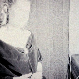 Here's a brief summary of the image: Woman in vintage dress sits near patterned curtains.