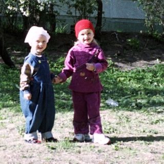 Two children stand near flowering trees and a building.