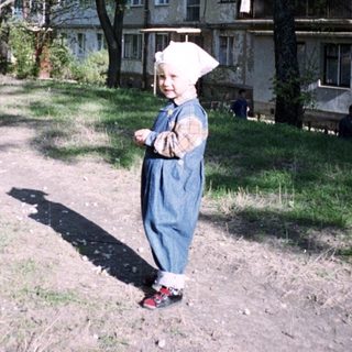 Here's a brief summary of the image: Child stands in a dirt path near an apartment building.