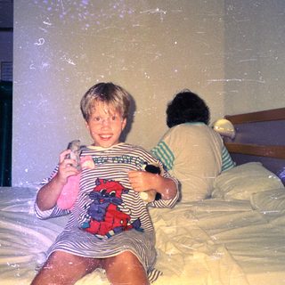 Child sits on a bed, holding toys, with another person behind.