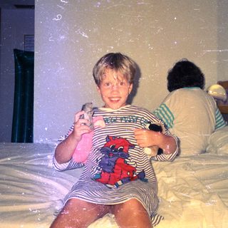 Child sits on a bed, holding toys, with another person behind.
