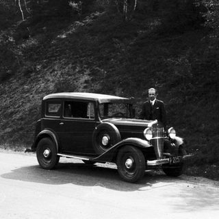 Man stands next to a vintage car on a road.