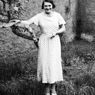 Woman in dress stands outdoors, posing near a tree.