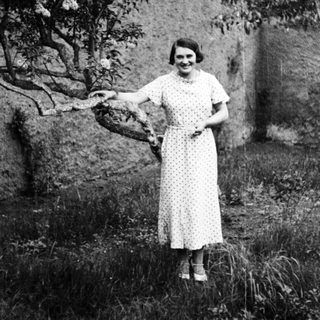 Woman in dress stands outdoors, posing near a tree.