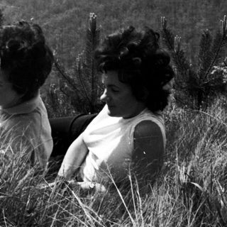 Three women seated in tall grass with a hilly landscape.