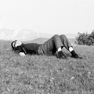Man reclines in a grassy field, landscape visible behind.