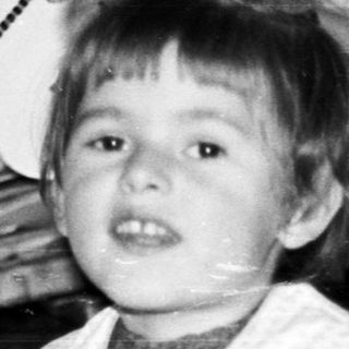Black and white photo of a child seated near a chair.