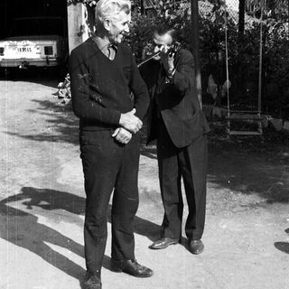Two men converse outdoors, one holding a camera.