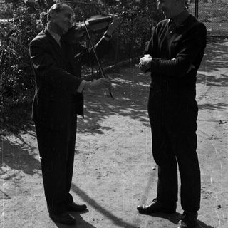 Two men outdoors; one presents a violin to the other.