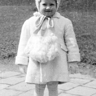 Here's a brief summary of the image: Young child in a coat and hat stands outside.