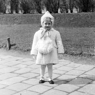 Here's a brief summary of the image: Young child in a coat and hat stands outside.