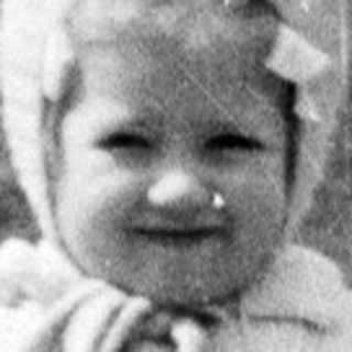 Here's a brief summary of the image: Young child in a coat and hat stands outside.