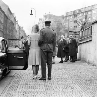 Military officer escorts women near a brick wall.