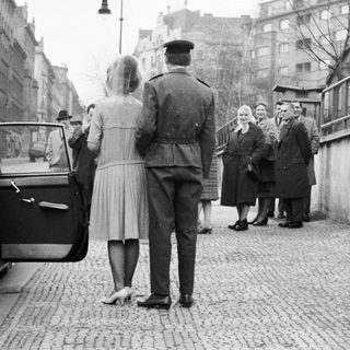 Military officer escorts women near a brick wall.