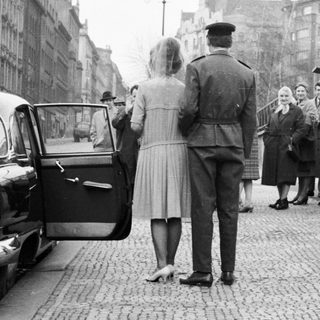 Military officer escorts women near a brick wall.