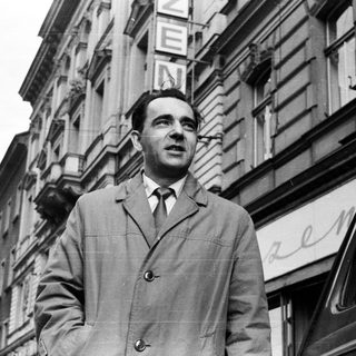 Man in coat stands outside a building in black and white.