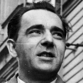 Man in coat stands outside a building in black and white.