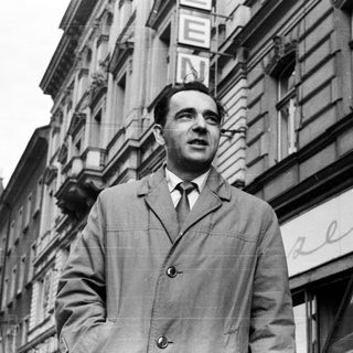 Man in coat stands outside a building in black and white.