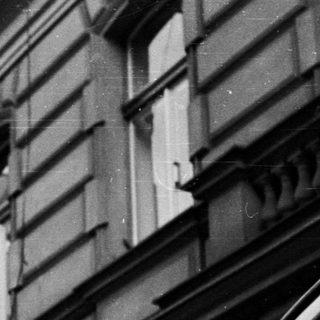 Man in coat stands outside a building in black and white.