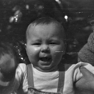 Baby in overalls cries, being held by an adult.