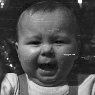 Baby in overalls cries, being held by an adult.