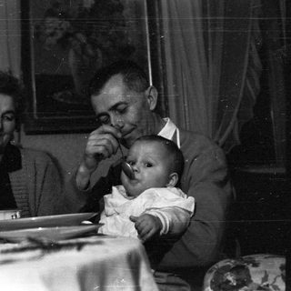 Man feeding a baby, with a woman seated nearby.