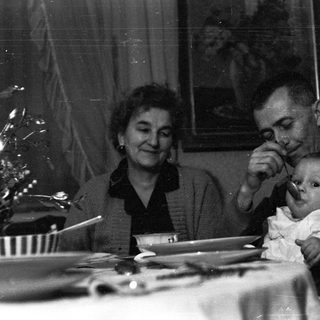 Man feeding a baby, with a woman seated nearby.