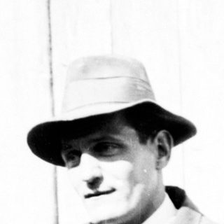 Man in suit holding flowers stands near a wooden building.