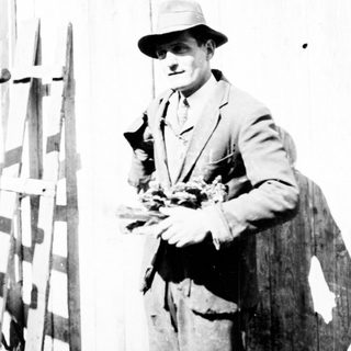 Man in suit holding flowers stands near a wooden building.