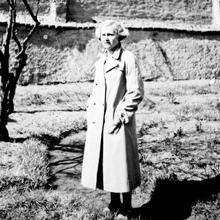 Woman in coat stands near a wall and tree.