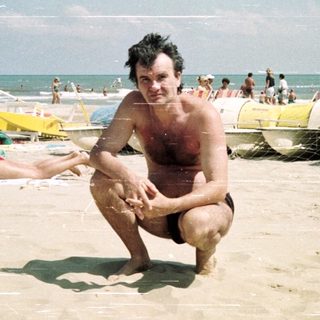 Man squats on a crowded beach with lifeguard equipment.
