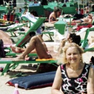 Woman sits on a beach filled with sunbathers and umbrellas.