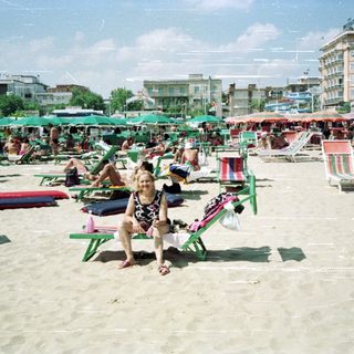 Woman sits on a beach filled with sunbathers and umbrellas.
