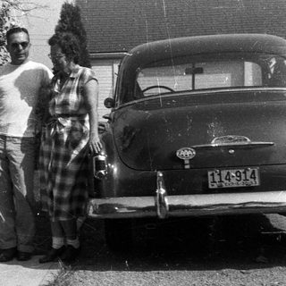 Three people stand near a vintage car and house.
